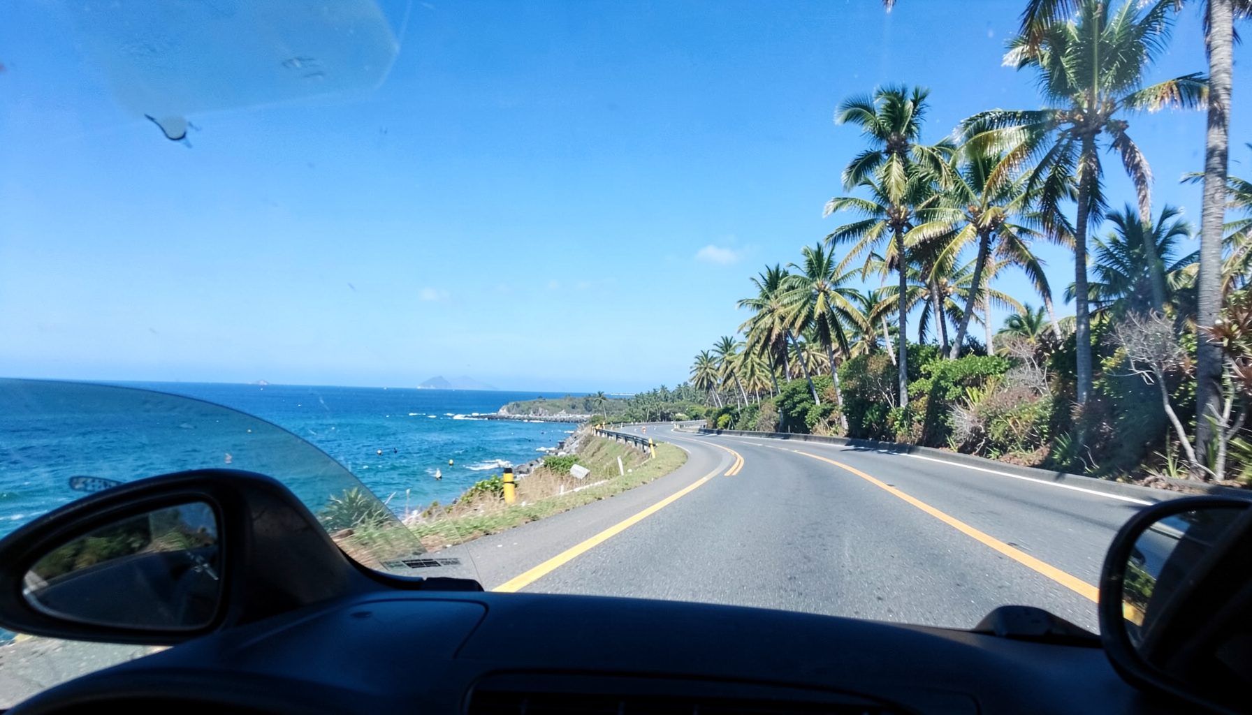 A winding coastal road with open car windows, palm trees and blue sky stretching ahead