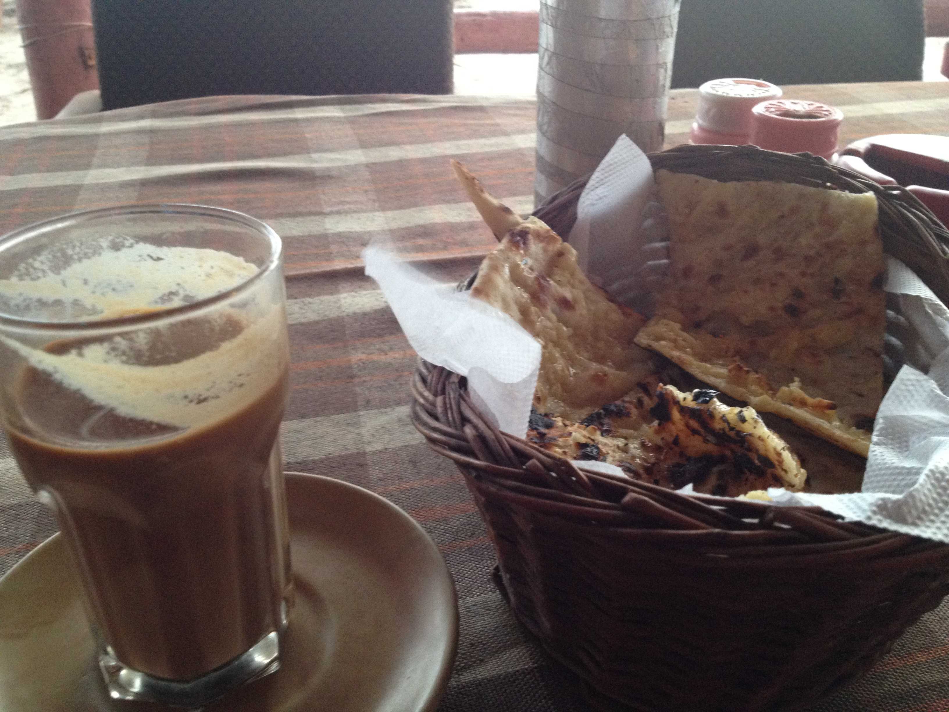 Glass of milky tea on a saucer next to a basket of flatbread on a checkered tablecloth