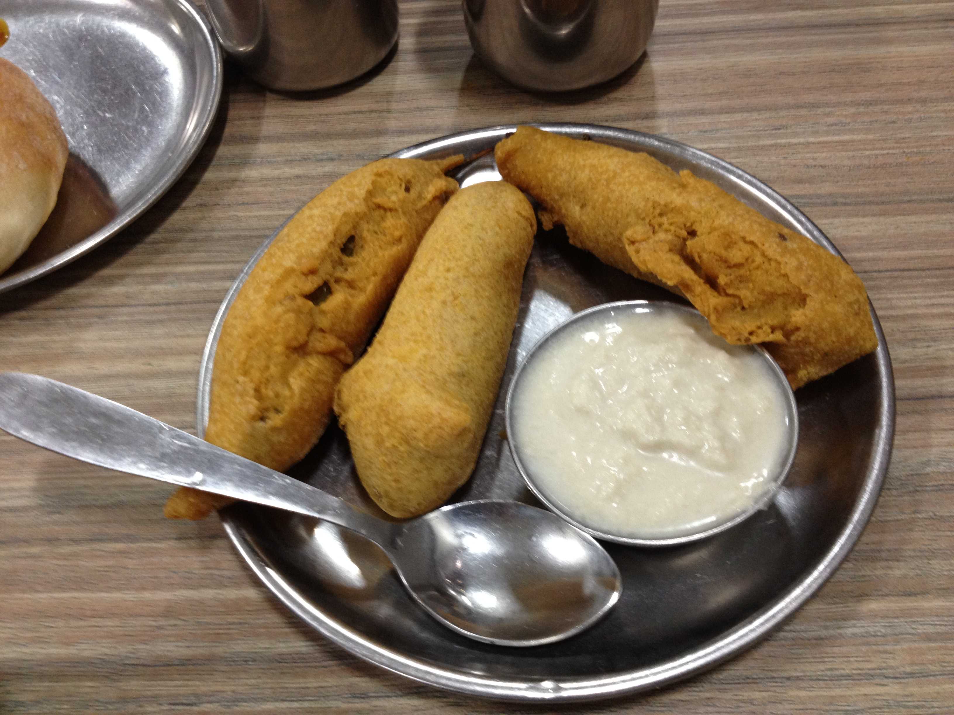 Three fried snack rolls on a metal plate with a bowl of white chutney and spoons, on a wooden table