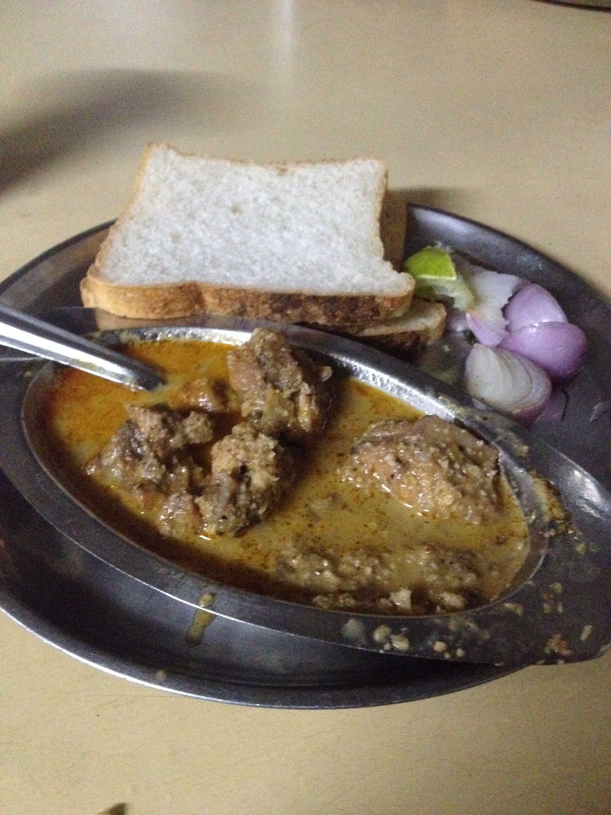 A metal plate with meat curry in rich gravy, bread slices, onion, and lime wedge on a table, warm brown and yellow tones