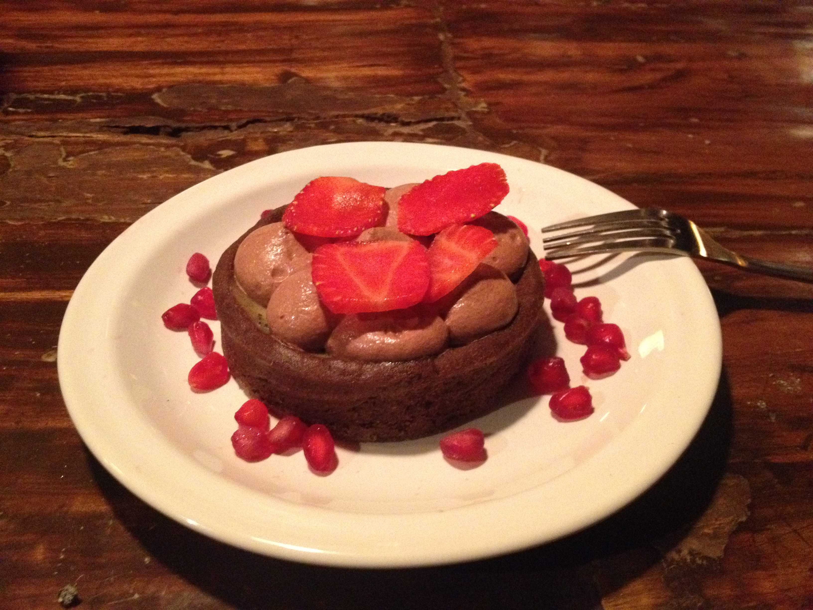 A round chocolate dessert topped with strawberries and pomegranate seeds on a white plate with a fork, on a dark wooden table